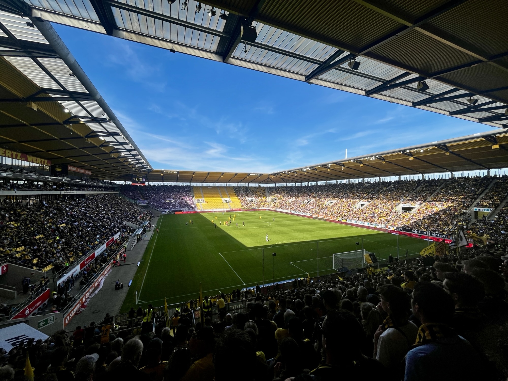 Weitwinkelaufnahme eines sonnigen Fußballspiels: Blick von einer überdachten Tribüne auf ein vollbesetztes Stadion (Tivoli Aachen) mit grünem Spielfeld und aktiven Spielern.