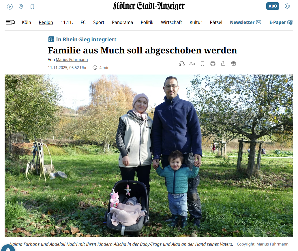 Das Bild zeigt eine Familie, die auf einer grünen Wiese im Freien steht. Im Vordergrund sitzt ein Baby in einer Babyschale mit einem Stofftier daran befestigt. Links daneben steht eine Frau mit heller Jacke, rechts ein Mann in dunkler Jacke, der ein kleines Kind an der Hand hält. Die Gesichter der Personen sind verpixelt. Im Hintergrund sind Bäume und ein Gartenschlauch, sowie weitere Menschen auf der Wiese zu sehen. Die Szene wirkt herbstlich, mit buntem Laub an den Bäumen und Sonne im Hintergrund