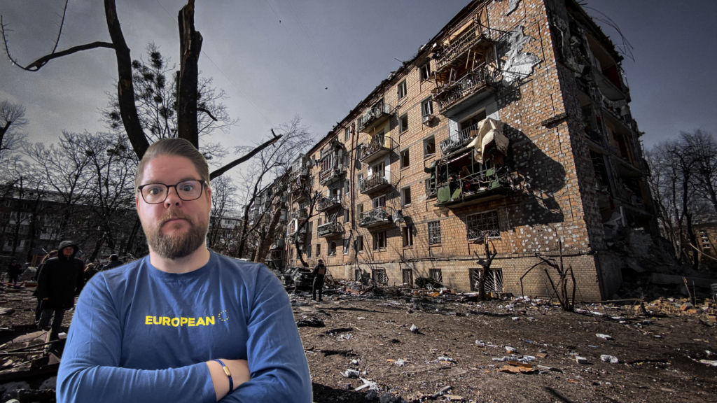 Mann mit Brille und blauem „European“-Shirt vor einem schwer beschädigten Wohnhaus in Kiew, Ukraine; im Vordergrund liegen Trümmer, im Hintergrund stehen Menschen zwischen zerstörten Bäumen und Gebäuden.