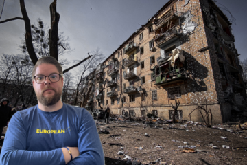Mann mit Brille und blauem „European“-Shirt vor einem schwer beschädigten Wohnhaus in Kiew, Ukraine; im Vordergrund liegen Trümmer, im Hintergrund stehen Menschen zwischen zerstörten Bäumen und Gebäuden.